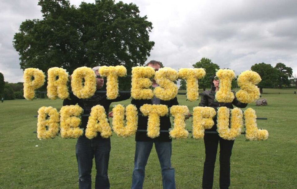 three people holding words made of yellow flowers saying protest is beautiful