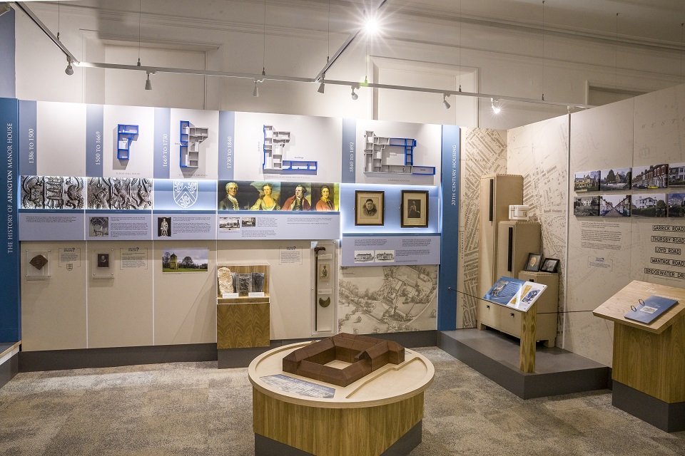 Gallery room with objects in cases and text panels on the walls. A central circular plinth with a wooden build a church interactive.