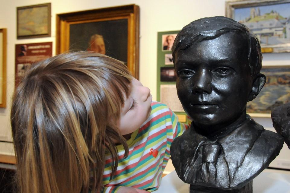 Image of a child with long hair with face turned away from the viewer looking up at a sculpture in dark bronze affect on a plianth.