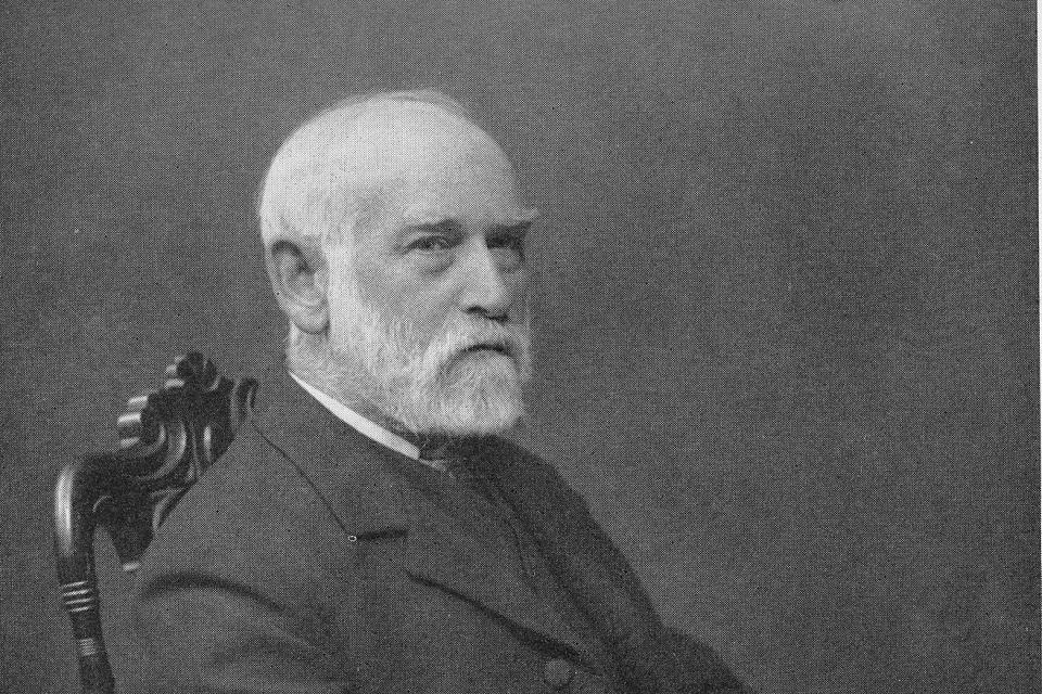 Black and white photograph showing a head and shoulders picture of an old man sitting in a chair. He has white hair and white facial hair, and is smartly dressed in a suit with a shirt, jacket, and tie.