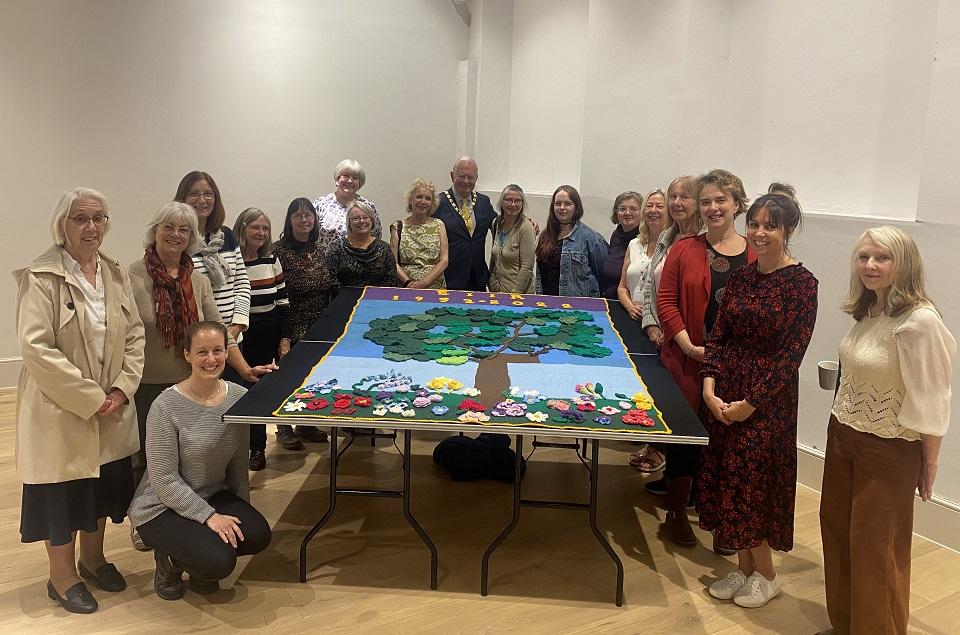 Group of 24 people suprrounding a large knitted colourful wool blanket. All people face the amera