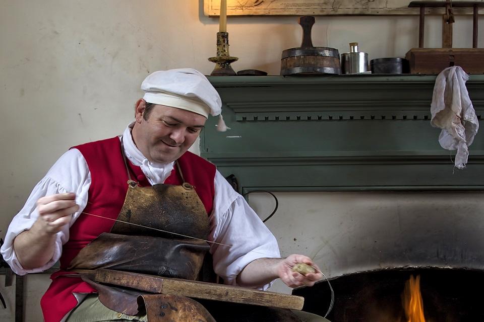 Colour photograph of a man in 1700 costume sitting in front of a fire making shoes. He rests his feet on a box of tools.