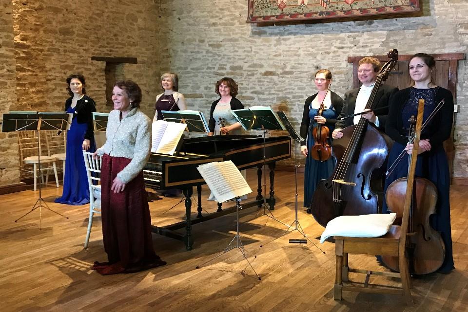 Seven people in a room with a white stone wall and wooden floorboards. They are all standing around a piano some holding violins, a cello and double bass.