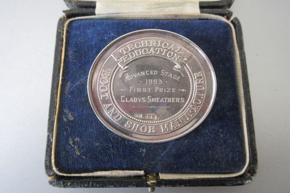 Silver first prize boot and shoe medal presented to Gladys Smeathers in 1953. A photo looking down on a square velvet lined open box containing a round silver medal with an image in scribed.