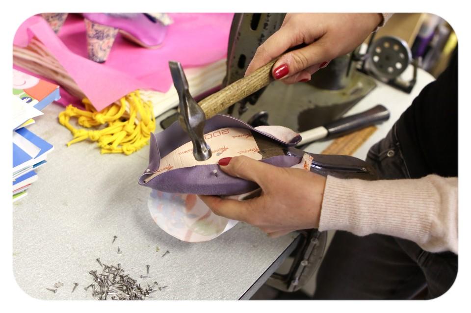 Photo looking down on a pair of hands shoemaking. The hands hold a yello foot shaped last and wood hammer with metal head..