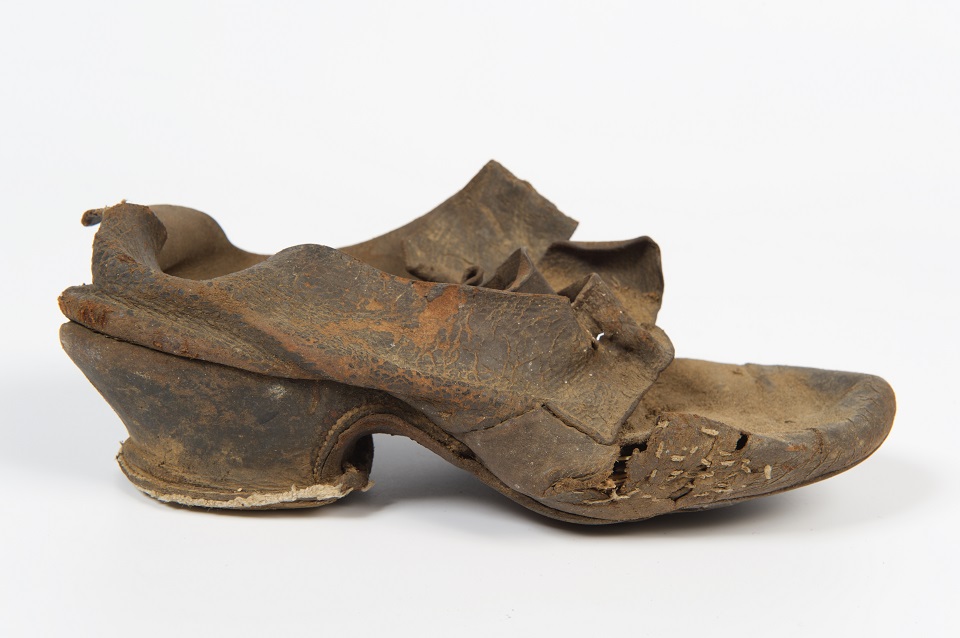 Women's black leather buckle latchet shoe, about 1750. It was found concealed in the flue above one of the ovens with a pair of shoes and a single shoe wrapped in a piece of brown paper, together with a leather gaiter and a wooden oven peel.