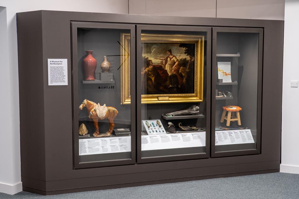 Image looks into a glass case containing a large painting in the centre with ceramic horse to the left, folssils in the centre and an animal stool.