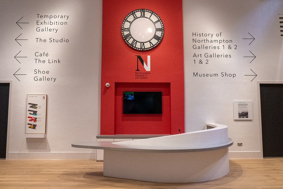 White desk with red wall behind displaying gallery directional signage and a clock.