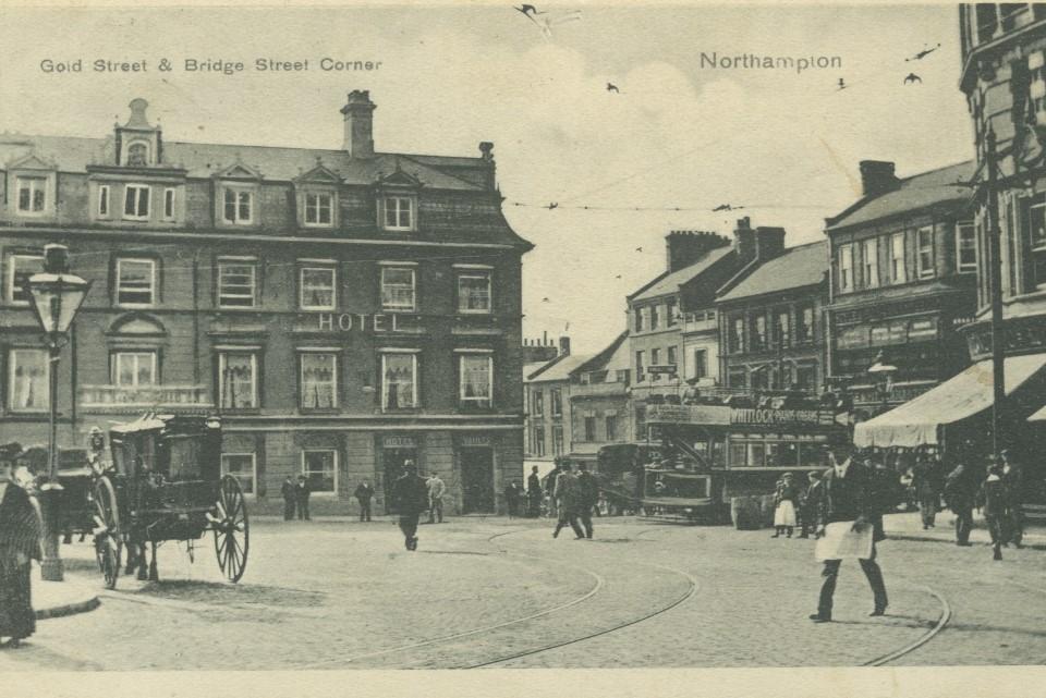 Sepia coloured photographic image depicting buildings at the back and people and horses in a street in front.
