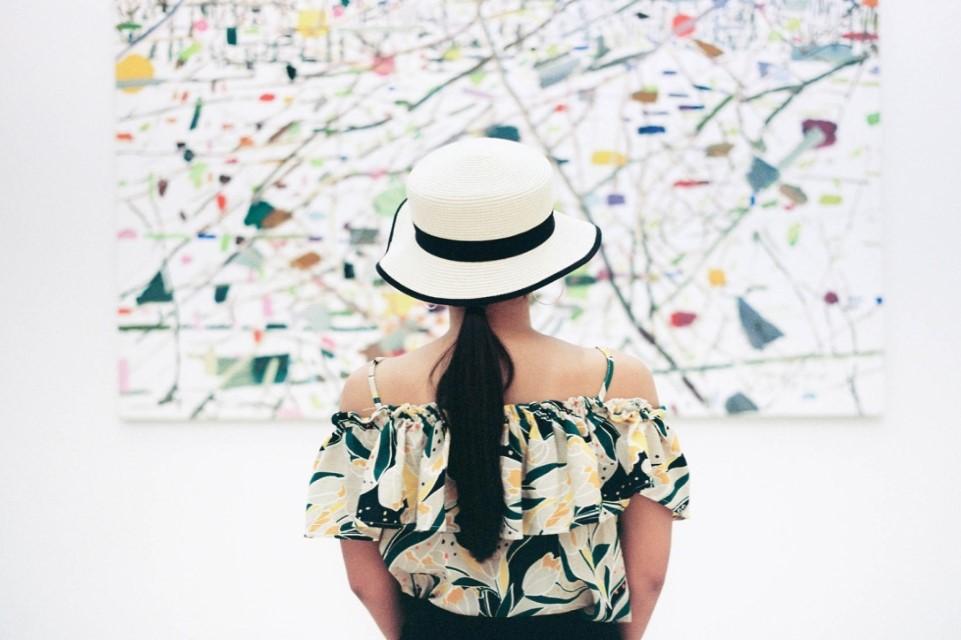 Photo of woman in white hat facing away from the camera looking at a pale abstracted art work haning in a white wall.