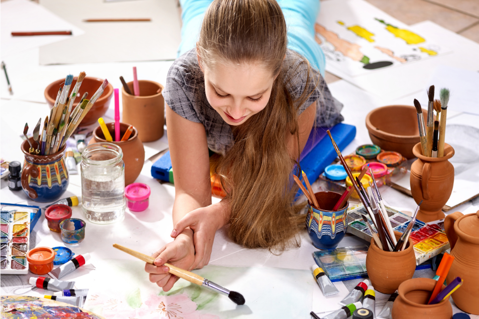 Photograph looking down on a child painting surrounded by paint and pots to the left and right