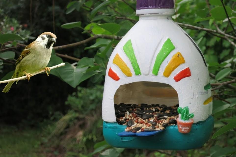 Bttle shaped white bird-feeder with rainbow markings and seed spilling from centre area. Hanging in green background with yellow and green and blue bird sitting to the left of the photograph.