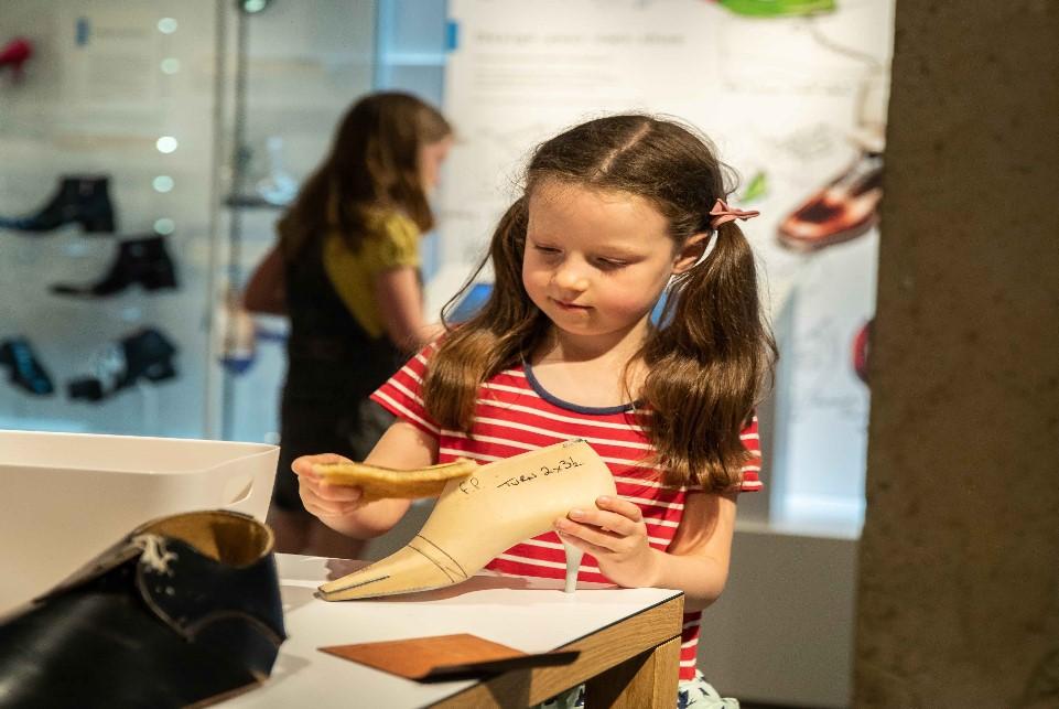 Child in red dress with bunches standing in front of cases of shoes. She holds a shoe last.