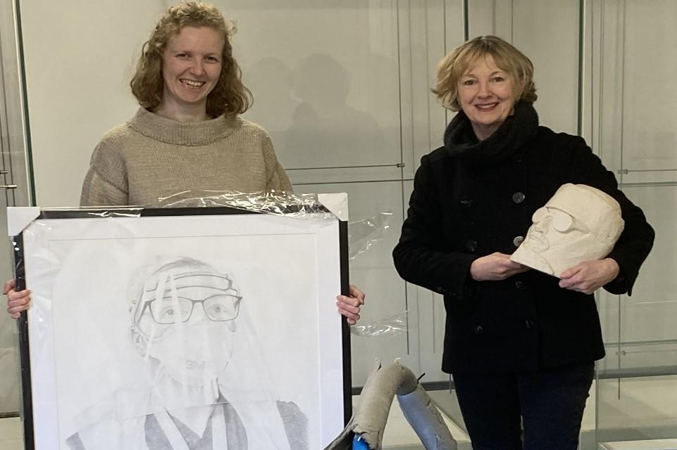 Image depicting two women standing next to each other looking at the camera. The woman on the right holds a sculptured head artwork and the woman on the right holds a pencil portrait.