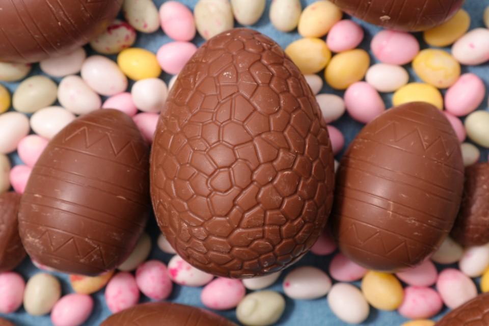 One large brown chocolate egg surrounded by two smaller eggs either side. The background is of pink, yellow and white round sweets