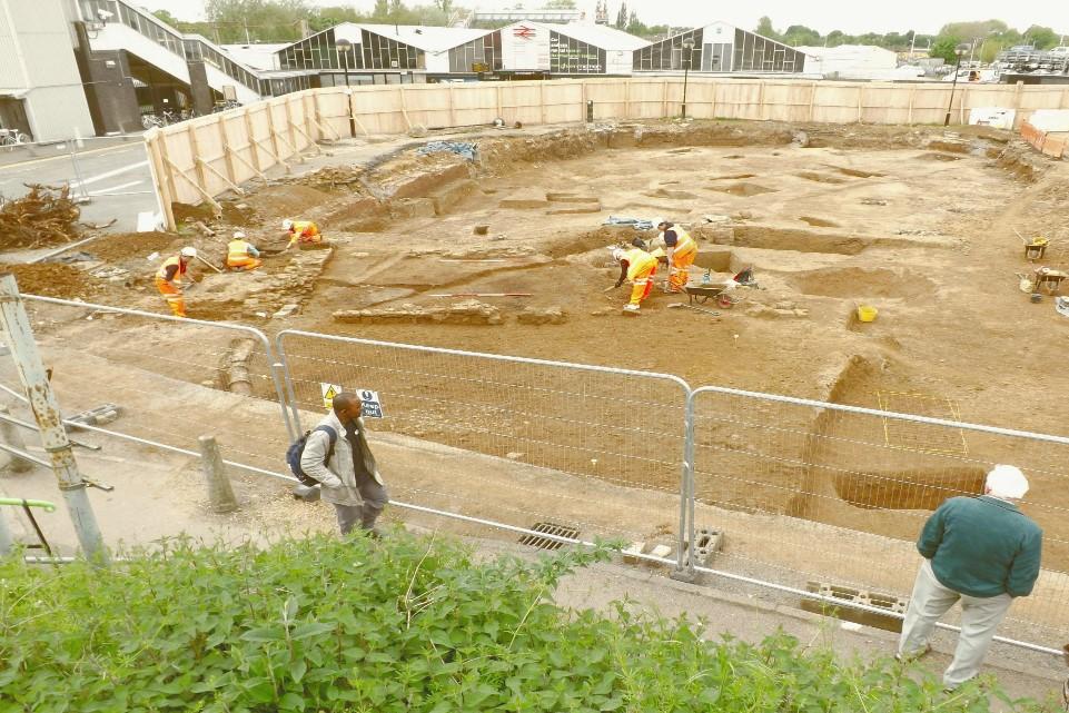 Phorograph. Soil underneath the surface is exposed in th photograhk as part of an archeological excavation. There are a number of small human figures working on the site. The imaf=ge is framed by green grass at the front and houses at the back.