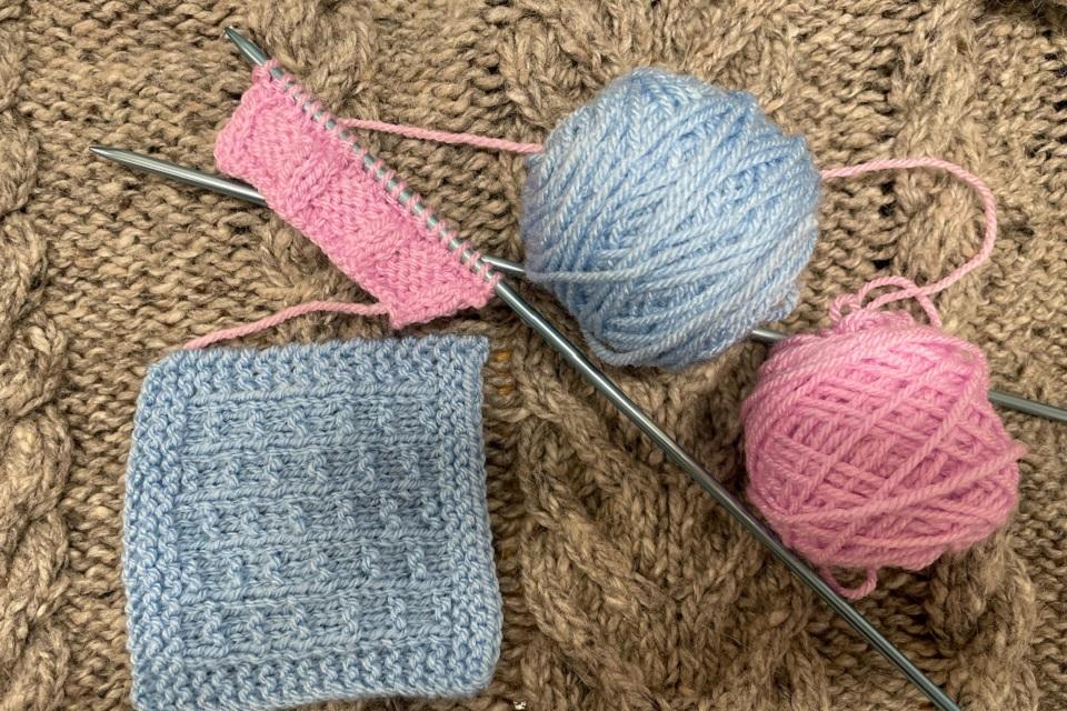 A photo showing a square of blue knitting, two long needles and two pink and blue half unwound balls of wool.