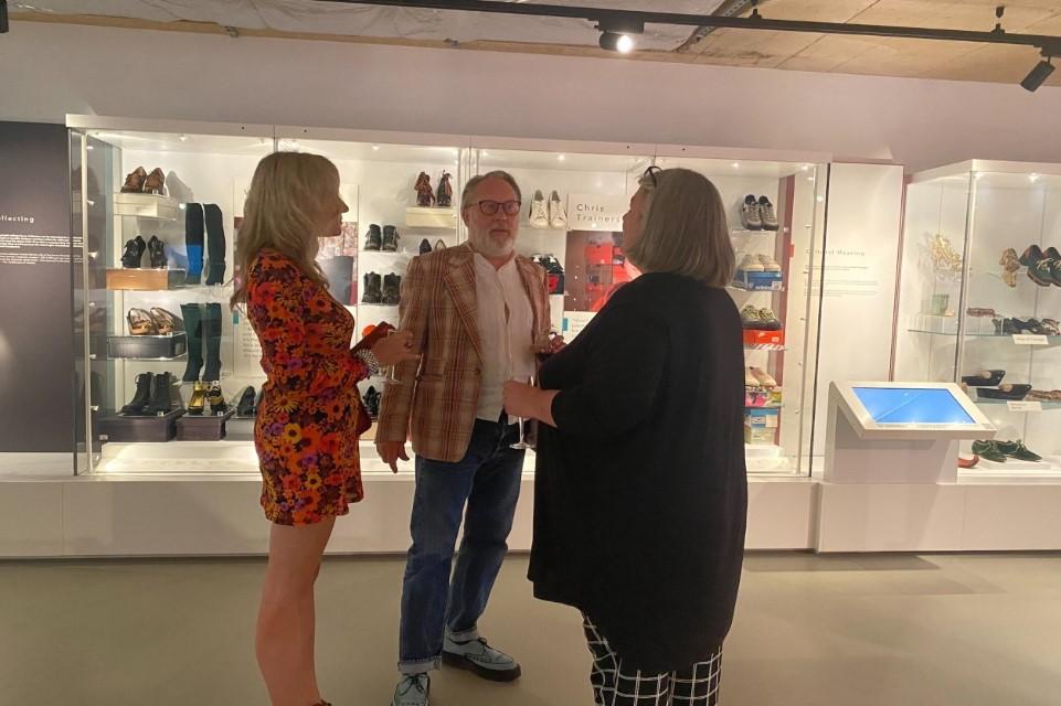 Three people standing in  a museum gallery space surrounded by shoes in cases. The central figure is Jim Moir aka Vic Reeves