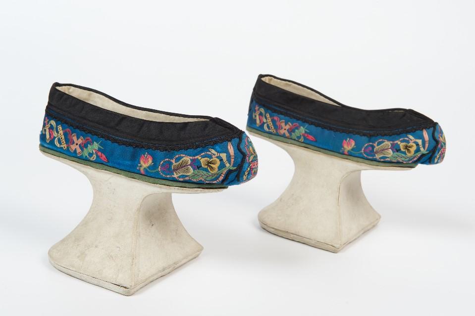 A pair of matching shoes pointing to the left. The shoes are on white stilts with narrow bases and the shoes are embriodered blue and gree patterns