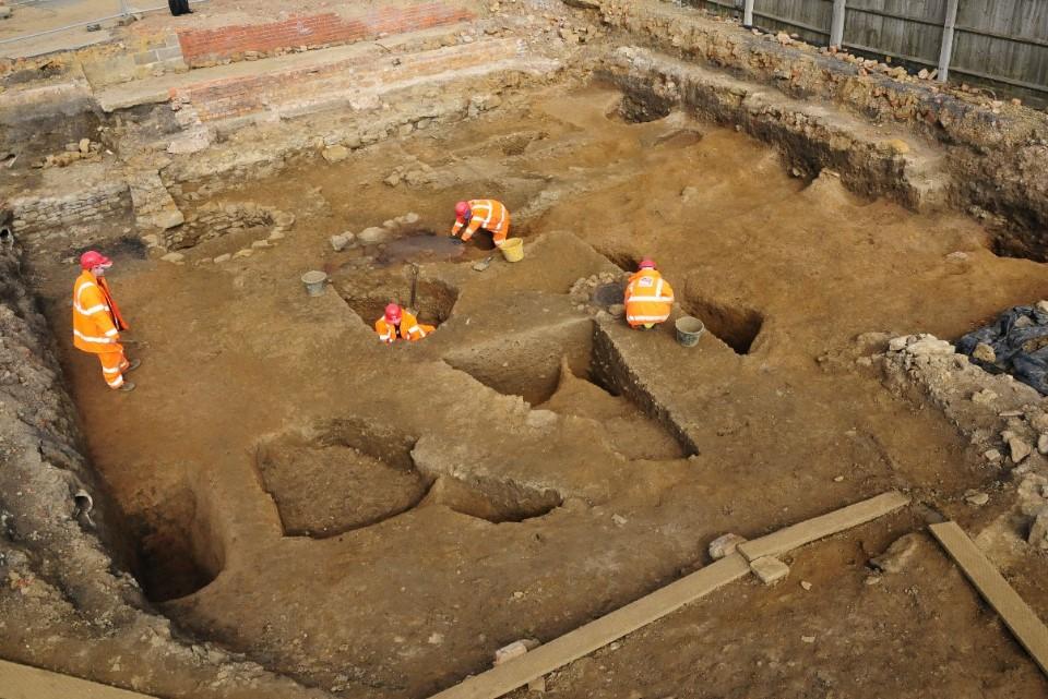 Photograph from above looking down on an archaeological excavation. Orange-brown soil showss various linear features and 2 crouched figures in orange digging.