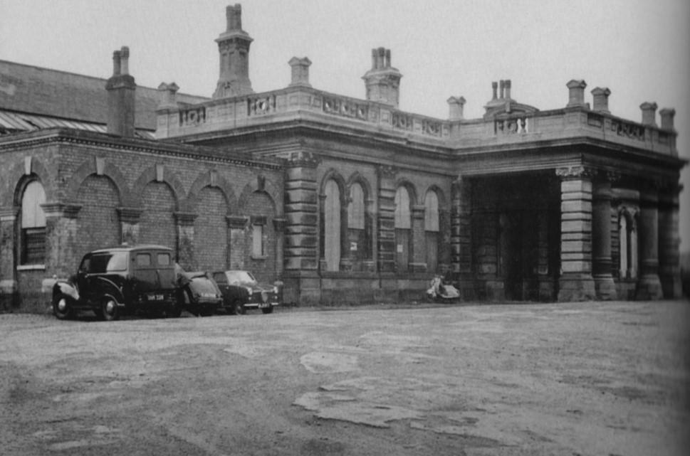 Black and white photograph showing a single story building with chimneys, decorative collumned porch and crenellated roof.