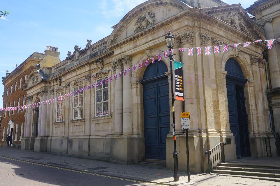 Image of light stone building with high entrance and classical design with blue sky behnd and street at the front.