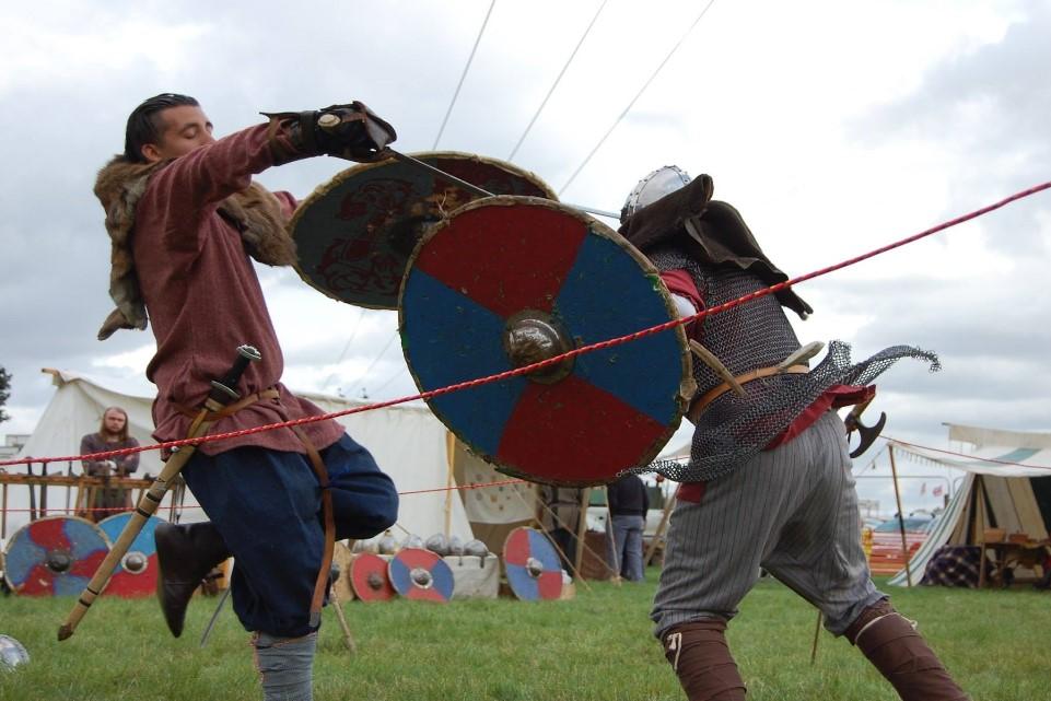 Image of two men fighting holding sheilds and swiords. A photo with tent in the background.