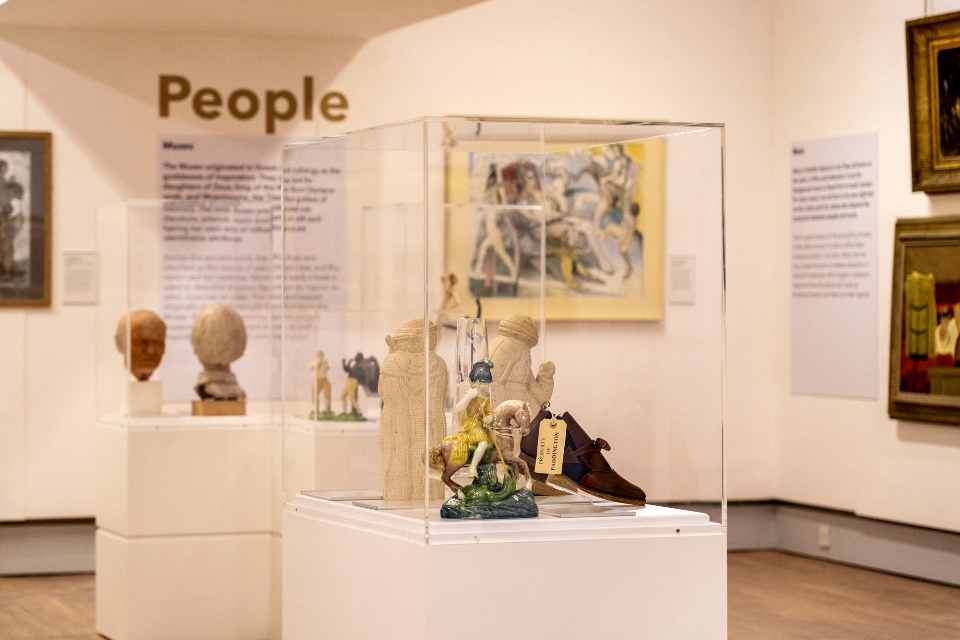 White plinths with objects on in cases and paintings on wall in background.