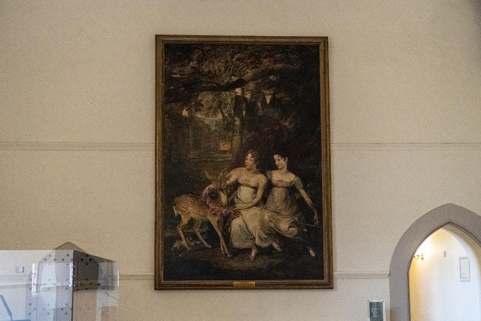 Large rectangular oil painting hanging on a wall depicting two women in long dresses and a deer.