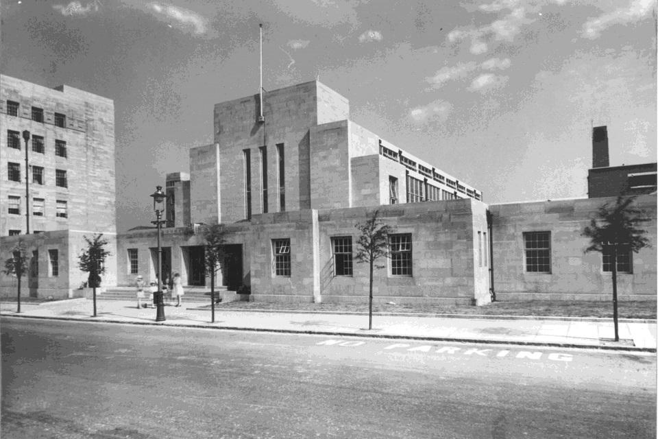 Black and white photograph depicting high rise square buildings viewed accross a road.