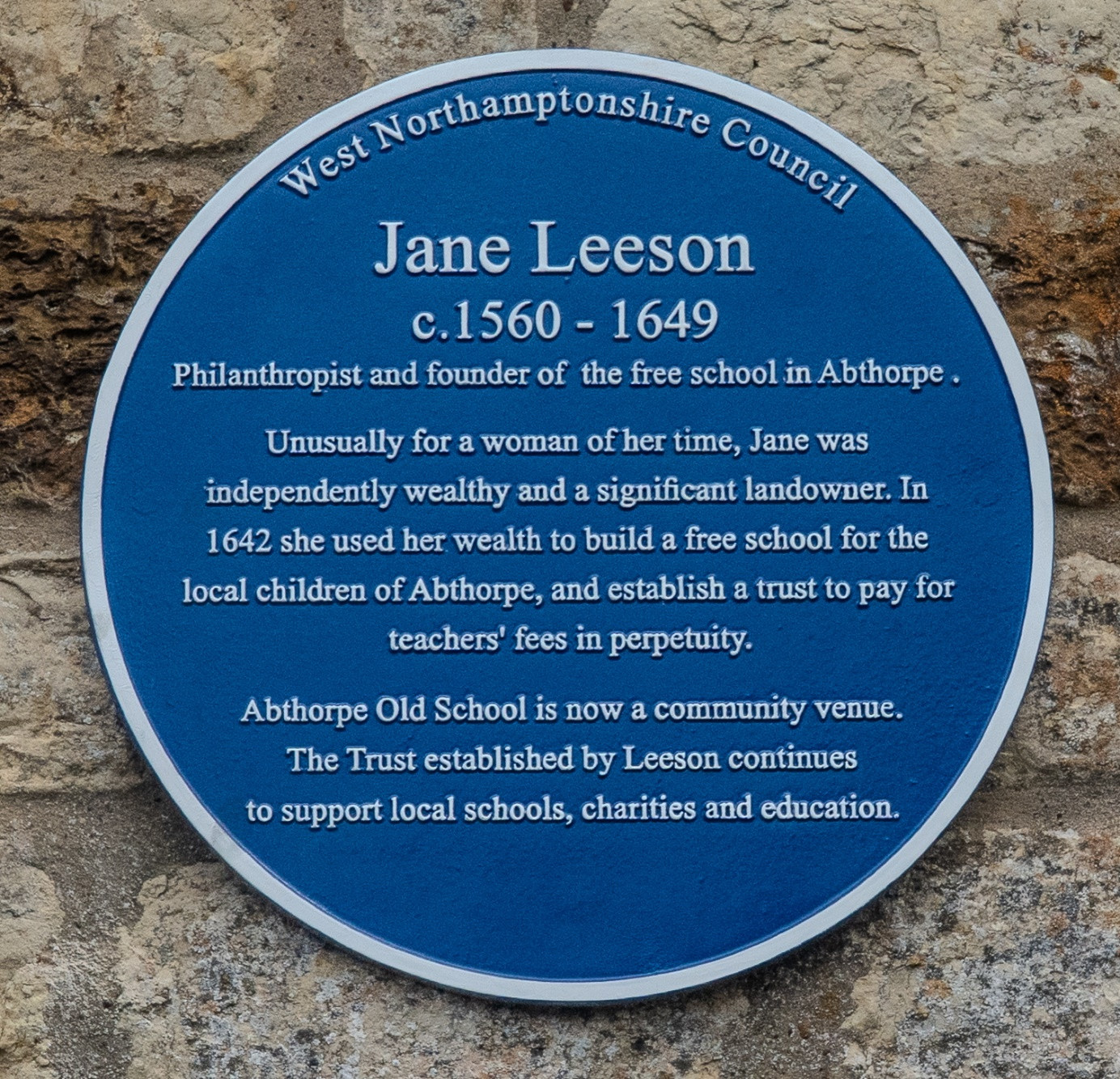 A blue circular plaque on a stone wall commemorating Jane Leeson, noting her role as a philanthropist and founder of the free school in Abthorpe, with text describing her contributions and legacy.