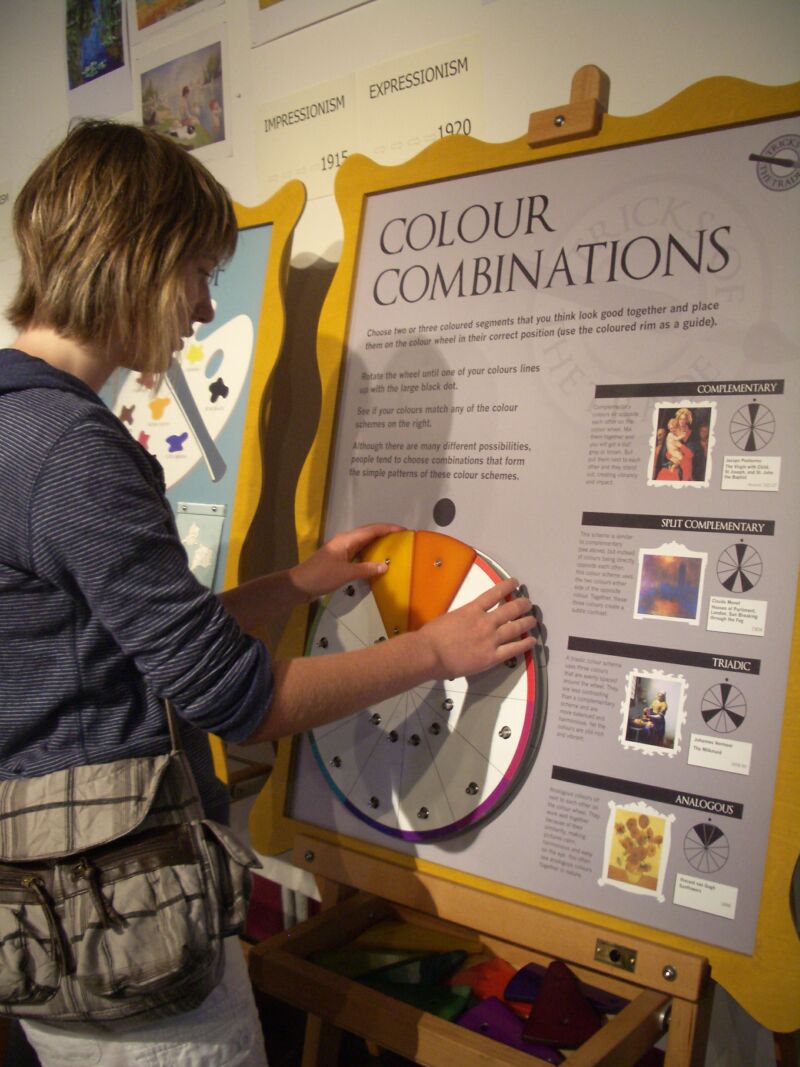 A person interacts with a hands-on museum exhibit titled “Colour Combinations,” rotating segments on a large colour wheel mounted on a display board. The board includes instructions and examples of colour schemes such as complementary, split complementary, triadic, and analogous. Additional artwork and a timeline labeled “Impressionism” and “Expressionism” are visible in the background, while coloured pieces are stored in a tray below the wheel.