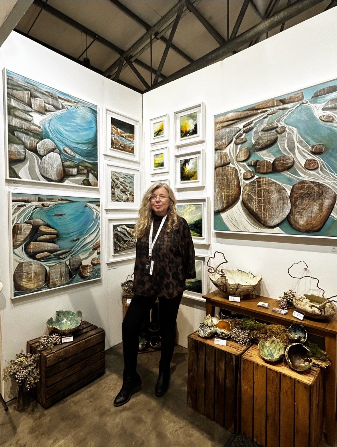 Person standing in an art gallery surrounded by framed coastal landscape paintings and ceramic pieces displayed on wooden crates.