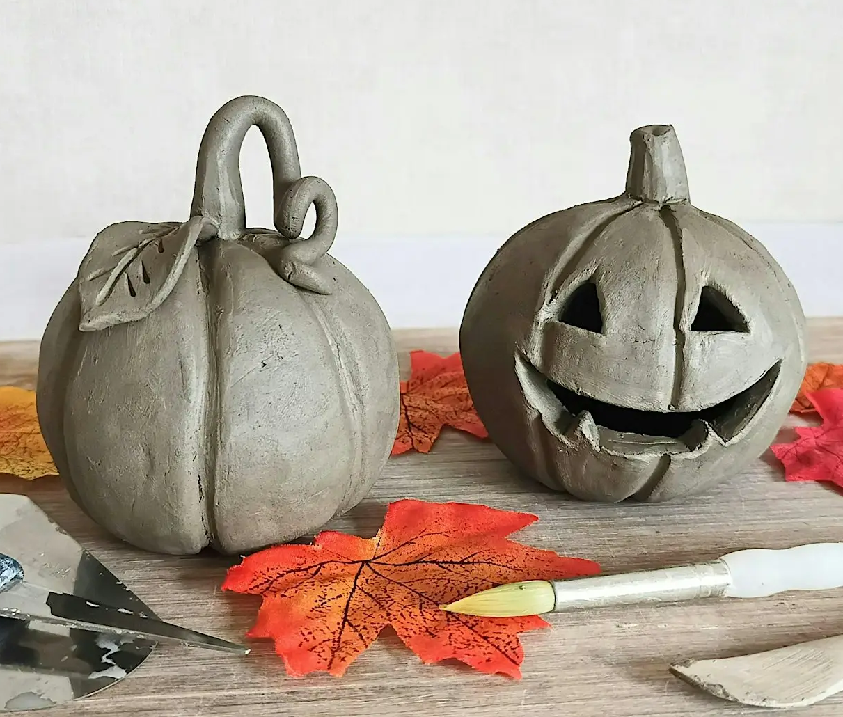 pumpkins Two clay pumpkins, one carved as a jack-o'-lantern, displayed with autumn leaves and pottery tools.
